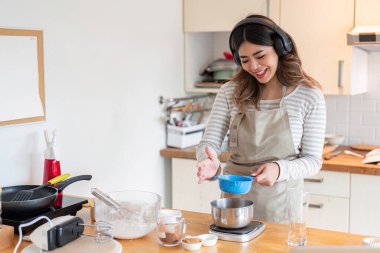 Önlük takan gülümseyen bir kadın mutfak terazisinde malzemelerini bir kaseye yerleştirir. Pişirme hazırlıklarını coşkuyla gösterir..