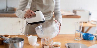 Önlüklü bir kadın ahşap mutfak masasında duruyor, tatlı hazırlamak için cam kasedeki malzemeleri karıştırıcı ile karıştırıyor..