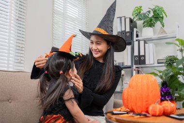 Bir anne, Cadılar Bayramı 'nı evde kutlarken kızlarının cadı şapkasını sevgiyle düzeltir. Etrafı şenlikli süslemeler ve balkabaklarıyla çevrilidir..