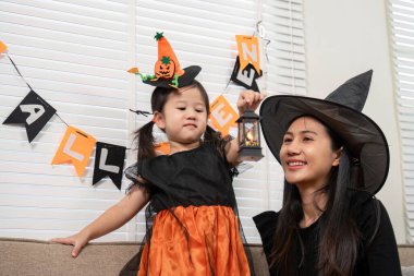 Cadı şapkalı bir anne ve kız Cadılar Bayramı 'nı evde kutlarken fener tutuyorlar. Etrafı şenlikli süslemeler ve afişlerle çevrili.