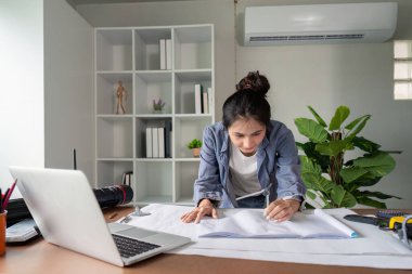 Odaklanmış bir mühendis, modern ofis ortamında ofis malzemeleri ve bir dizüstü bilgisayarla çevrili planları masasında analiz eder..