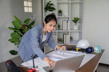 Bir mühendis, modern bir çalışma alanındaki araçlar ve ofis malzemeleriyle çevrili planlar üzerinde çalışmak için dizüstü bilgisayar kullanır..