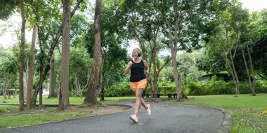 Yeşil bir parkta kaldırımlı bir yolda koşan yaşlı bir kadın, fitness ve sağlıklı bir yaşam tarzını teşvik ediyor..