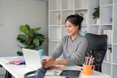 Modern bir ofiste renk paletleri ve tasarım araçlarıyla çevrili bir dizüstü bilgisayarda yazı yazan bir grafik tasarımcısı yaratıcılığı sergiliyor..