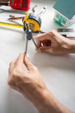 Bir mühendis, bir masanın üzerinde kesin ölçümler için pusula kullanır, yanında model bir ev ve modern bir ofiste mezura bulunur..