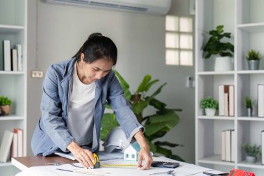 Kot ceketli bir kadın mühendis, modern bir çalışma alanındaki ofis malzemeleriyle çevrili bir model evin yanında bir bant ile ölçer..