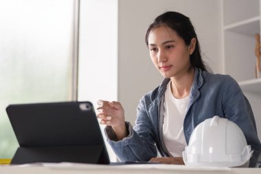 Kot ceketli bir kadın mühendis, modern bir ofiste proje yönetimi için tablet kullanıyor, yanında dijital aletlere odaklanan bir kask var..