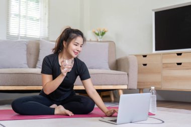 Siyah giysili bir kadın evdeki yoga seansı sırasında su molası veriyor. Sıcak bir oturma odasında dizüstü bilgisayarla oturuyor..