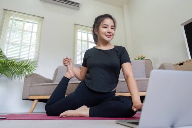 Siyah giysili bir kadın evdeki minder üzerinde yoga yapıyor, rahat bir oturma odasında doğal ışığı olan bir dizüstü bilgisayar kullanıyor..