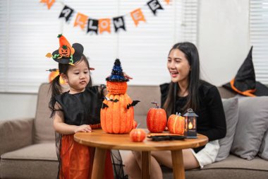 Asyalı bir anne ve kız Cadılar Bayramı balkabağı yapmaktan zevk alıyor, evde dekorasyonlarla neşeli ve neşeli bir atmosfer yaratıyor..