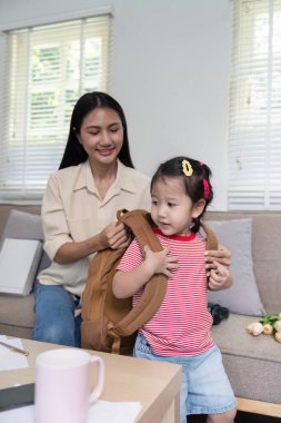 Bir anne, sabah rutini sırasında kızına sırt çantası organizasyonunda yardımcı olur, hazır olduğunu ve dikkatli bir yardım aldığını vurgular..