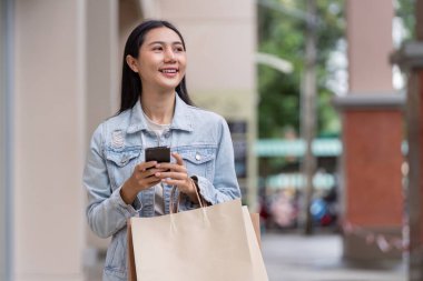 Neşeli bir alışverişçi akıllı telefonuyla iletişim kurar, alışveriş sonrası heyecanı olan bir alışveriş çantası taşır..