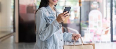 Modadan anlayan genç bir kadın akıllı telefonunu bir alışveriş merkezinde kullanıyor. İnternetteki satışlarla ilgilenmeye ve alışveriş deneyimini tarzıyla geliştirmeye hazır..