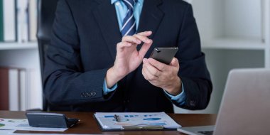 Takım elbiseli bir iş adamı, ofisinde dinamik bir çalışma ortamı sergileyerek, akıllı telefonunda güncelleme var mı diye kontrol ederek çoklu görev yapar..