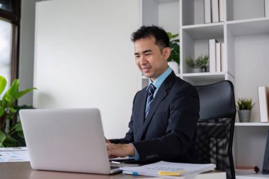 Ofisinde dizüstü bilgisayarla çalışırken gülümseyen bir işadamı, profesyonel bir ortamda pozitifliği ve bağlılığı temsil eder..