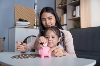 Asyalı anne ve kız evlerinde pembe bir domuz kumbarası kullanarak öğrenme ve kaynaşmayı teşvik ederek para tasarrufu faaliyetinde bulunuyorlar..