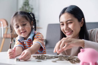 Asyalı anne ve kızı mutlu bir şekilde madeni paraları bir araya getirip, pembe domuzcuk bankasını kullanarak evde sevgi dolu bir finansal öğrenme deneyimini teşvik ediyorlar..