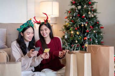 Lezbiyen Asyalı çift tatil ruhuyla online alışverişin keyfini çıkarıyorlar. Kredi kartıyla. Etrafı şenlikli süslemeler ve Noel ağacı ile çevrili..