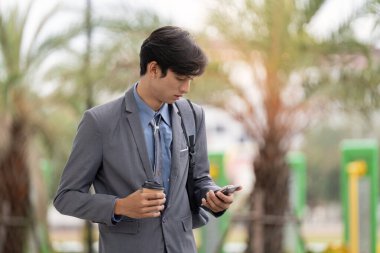 Şehir parkının yakınında kahve ve akıllı telefon hokkabazlığı yapan odaklanmış Asyalı bir adam, yoğun şehir yaşamını temsil ediyor..