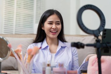 Asyalı kadın evde makyaj dersi kaydederken gülümsüyor. Güzellik ürünleri sergiliyor. Seyircilerini etkilemeye odaklanıyor..