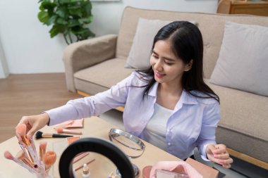 Genç Asyalı kadın makyaj fırçalarını ve aletlerini parlak bir oturma odasında bir masanın üzerinde düzenliyor ve makyaj seansına hazırlanıyor..