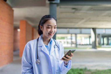 Açık havada akıllı telefon kullanan gülümseyen Asyalı kadın doktor, steteskoplu beyaz önlük giyiyor..