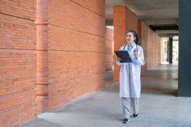Asyalı kadın doktor tuğla koridorda yürüyor, elinde bir dosya ve kahve var, profesyonel kararlılık gösteriyor..