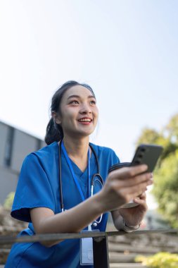 Asyalı hemşire neşeyle kahve ve akıllı telefon tutuyor, mavi önlük giyiyor ve açık havada steteskop takıyor..
