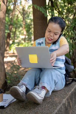 A woman with achondroplasia using a laptop and phone outdoors, showcasing remote work and technology. clipart