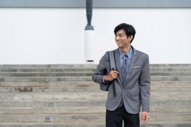 Asyalı işadamı sırt çantası taşırken gülümsüyor. Profesyonel bir ortamda rahat ve kendinden emin bir duruş sergiliyor..