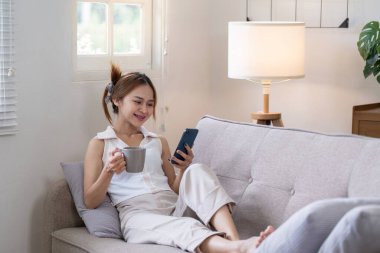 Genç bir kadın kanepede rahatlıyor, kahvesini yudumluyor ve rahat bir oturma odasında telefonunu kullanıyor..