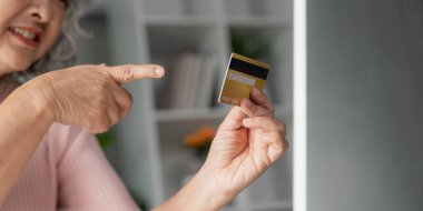 Hevesli, yaşlı bir kadın kredi kartını işaret ederek ev ofisinin rahatlığı içinde online alışveriş deneyiminin altını çiziyor..