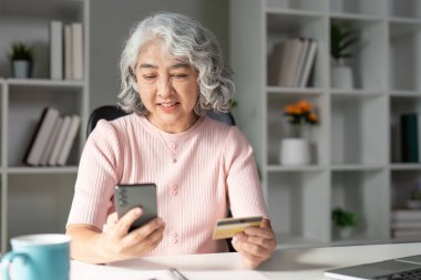 Yaşlı bir kadın elinde kredi kartı tutarken gülümsüyor ve akıllı telefonunu internet üzerinden alışveriş yapmak için kullanıyor..