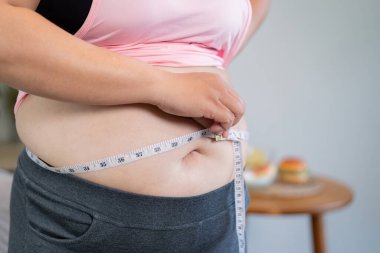 Pembe bluzlu bir kadın belini mezurayla ölçer. Hamburgerle yan yana gelir. Ağırlık yönetimi yolculuğundaki dengeyi koruma mücadelesini temsil eder..