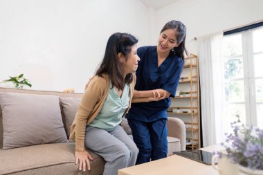 Bakıcı, yaşlı bir kadına fiziksel destek sağlarken aynı zamanda kanepeden kalkmasına yardım ederek sıcak bir ev ortamında güvenlik ve konfor sağlıyor..