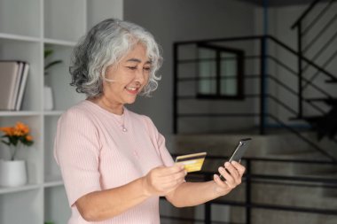 A happy senior woman enjoying her online shopping experience, holding a credit card and smartphone in a beautifully designed living space. clipart