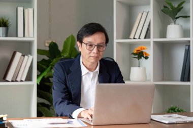 Üst düzey bir profesyonel gözlük takıyor, özenle dizüstü bilgisayarda çalışıyor, yeşilliklerle çevrili modern ofis ortamında raporları analiz ediyor..