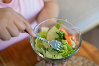Kilolu bir kadının eli renkli bir salataya çatal batırıyor ve evde daha sağlıklı bir yaşam tarzı ve kilo verme konusundaki kararlılığını vurguluyor..
