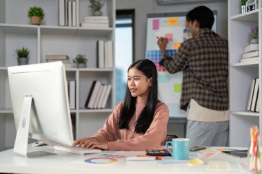 Canlı bir çalışma alanında bilgisayarda yazı yazan bir grafik tasarımcısı, bir meslektaşı dinamik bir tasarım projesine katkıda bulunarak bir ruh hali tahtasında renkli notlar düzenlerken,.