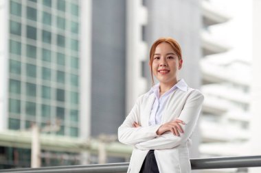 Beyaz ceketli kendine güvenen bir iş kadını kollarını kavuşturarak gururla durur. Gösterişli, çağdaş bir ofis yapısına gülümser..