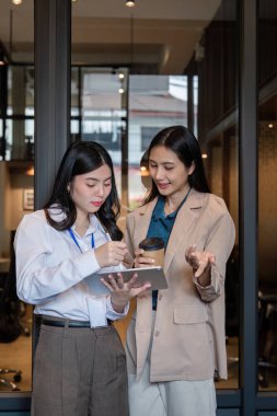 Üretken bir sohbete girişen iki profesyonel kadın, bir tanesi elinde kahve tutarken, şık bir ofis ortamında dijital tabletin proje bilgilerini gözden geçiriyor..