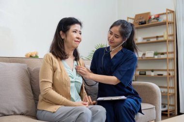 A caregiver attentively listens to the heartbeat of an elderly woman with a stethoscope, showcasing the importance of regular health assessments in a comfortable environment. clipart