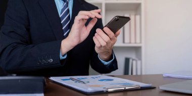 Odaklanmış bir işadamı, finansal raporları analiz ederken, görevleri yönetirken ve çağdaş bir ofis ortamında iletişimde kalırken akıllı telefonuyla etkileşim halindedir..