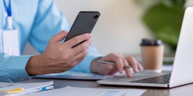 Bilgisayarda çalışırken akıllı telefon kullanan bir iş adamı, görevleri çağdaş ofis ortamında verimli bir şekilde yönetir.