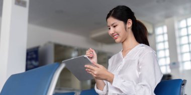 A cheerful woman enjoys using her tablet while seated in an airport lounge, eagerly ready for her upcoming travel experience. clipart