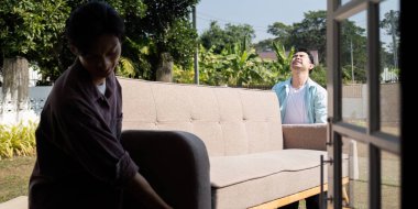 A joyful scene of two men, a couple, moving a spacious sofa into their house while sharing laughs, surrounded by greenery outside. clipart