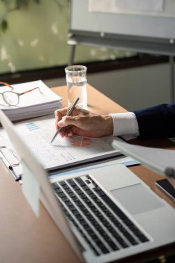 Finansal raporları inceleyen ve belgelerle ve dizüstü bilgisayarla çevrili şık bir ofiste kalemle notlar yazan kıdemli bir analist..