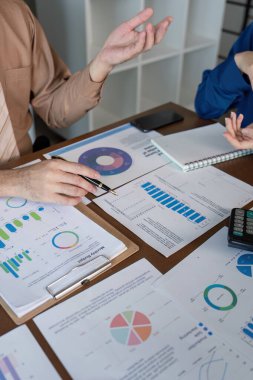 İki muhasebeci, modern bir ofis ortamında takım çalışması sergilerken finansal eğilimler, grafikler ve verileri analiz eden dinamik bir tartışmaya giriştiler..