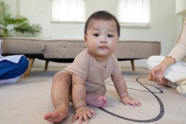 Meraklı bir bebek yerde oturuyor, merakla ebeveynlerine bakıyor, ilgili bir çocuk doktoru yakınlarda sıcak, davetkar bir yerde izliyor..