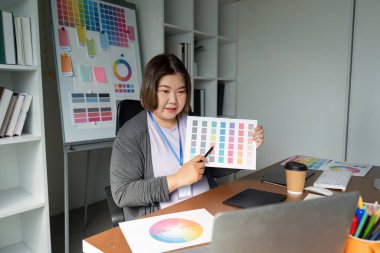 Grafik tasarımcısı, masasında otururken çağdaş bir ofis ortamında dizüstü bilgisayar ve tasarım materyalleri ile çeşitli renk paleti seçenekleri sunar.